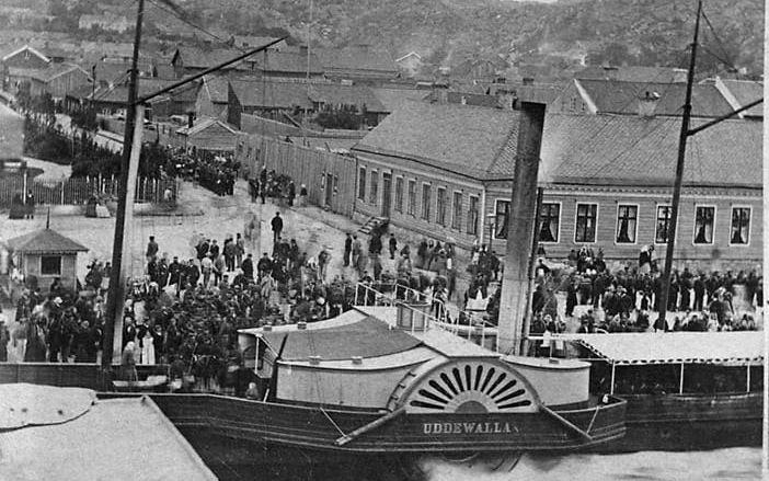 Trängsel runt hjulångaren ”Uddewalla” år 1865. Till vänster syns den minimala biljettkiosken. Vändbassängen, som anlades delvis för ”Uddewallas” skull, ligger på Västerbrons nuvarande plats. Ångfartyget ”Uddewalla” var en praktsensation när det år 1853 levererades från Motala verkstad. Med sina 60 hästkrafter var det Sveriges snabbaste fartyg. Sträckan mellan Göteborg och Uddevalla – som med tidigare förbindelser tagit dagar eller rentav veckor – tog plötsligt åtta timmar. Det ansågs av samtiden var chockartat snabbt. Det betraktades också som vådligt och dödsföraktande att gå i öppen sjö. När bilden togs var dock ”Uddewalla” redan på väg att bli omodernt. Inom några år skulle hjulångarna ha slagits ut av propellerfartyg. Fotografiet togs troligen i mitten av 1860-talet av Maria Lundbäck – en av Uddevallas första professionella fotografer. Hon använde troligen den då populära våtplåtsmetoden som gjorde det möjligt att ta detaljerade bilder, men som krävde framkallning inom 10-15 minuter. Några människor har rört sig under exponeringen och kan följaktligen anas som grå och spöklika dubbelfigurer på kajen.
